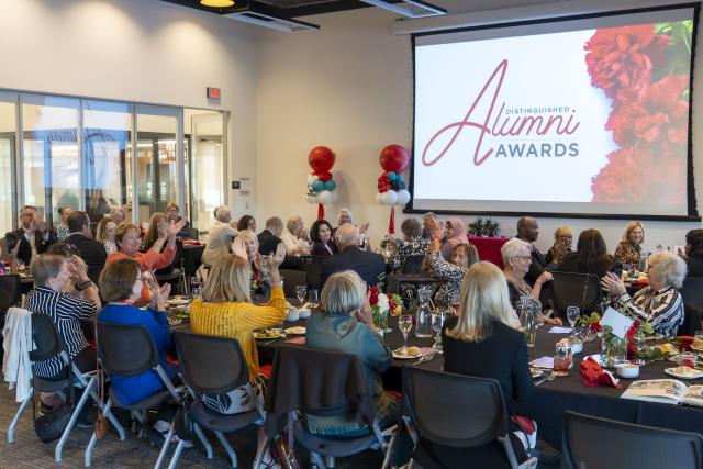 A room of people clapping and smiling by a projected screen that says "Alumni Awards"